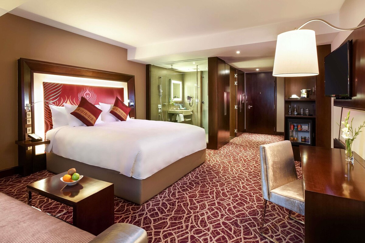 Habitación de hotel con una cama grande, una mesita de noche con fruta, una zona de estar con un sillón, un escritorio con lámpara y un armario.