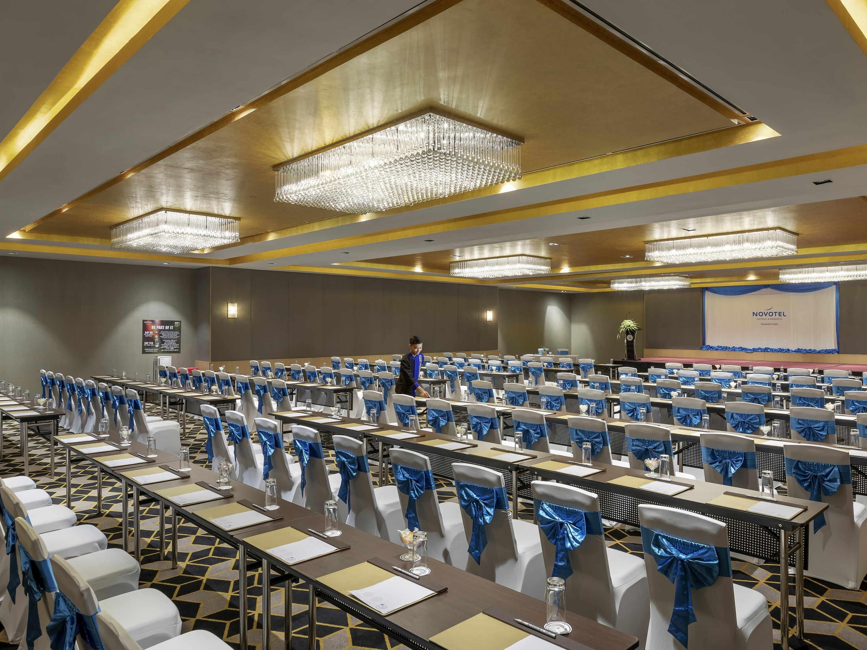 Un salón de conferencias con filas de mesas, sillas con cintas azules, candelabros y un escenario con una pantalla que muestra 'NOVOTEL'.
