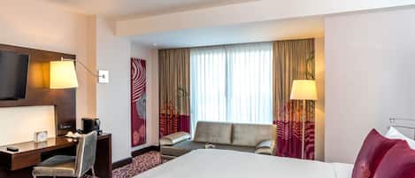A hotel room with a large bed, a desk with a chair, a TV, and a window with curtains.