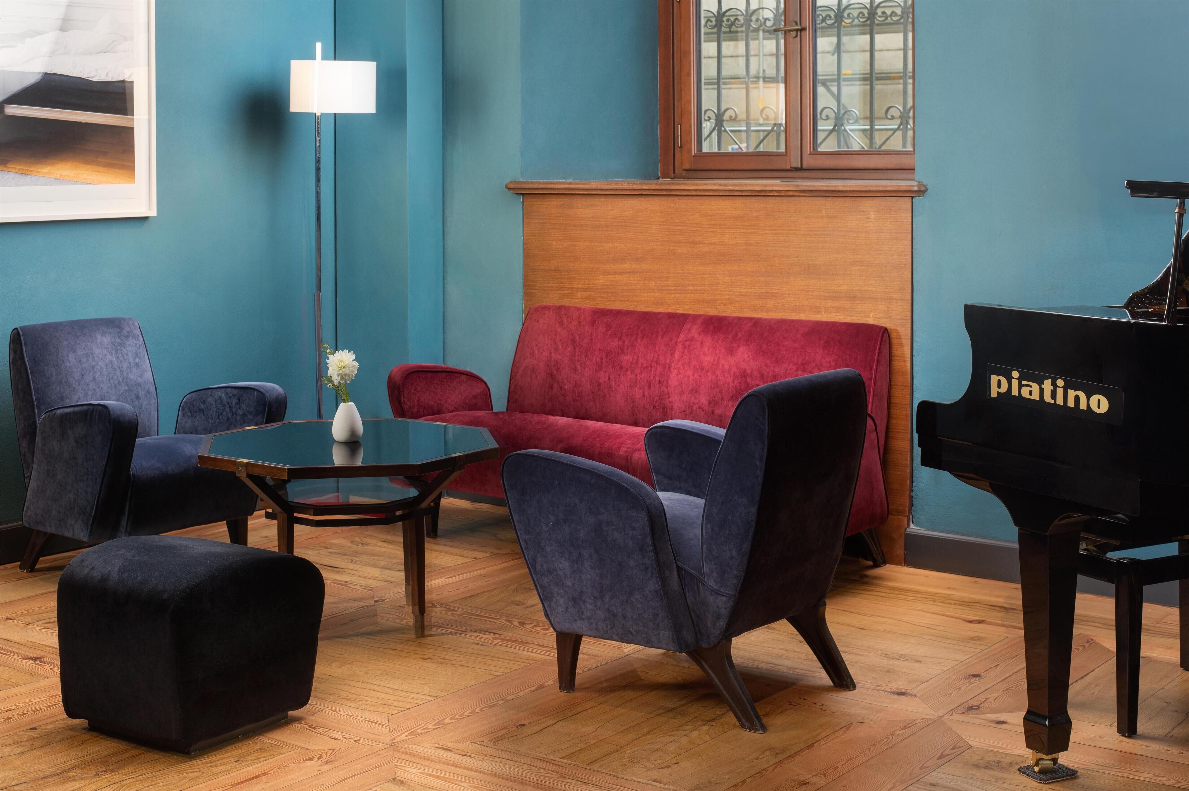 Une pièce avec un canapé rouge, deux fauteuils bleus, un pouf noir, une table basse en verre, un lampadaire et un piano à queue portant l’inscription « Piantino ».