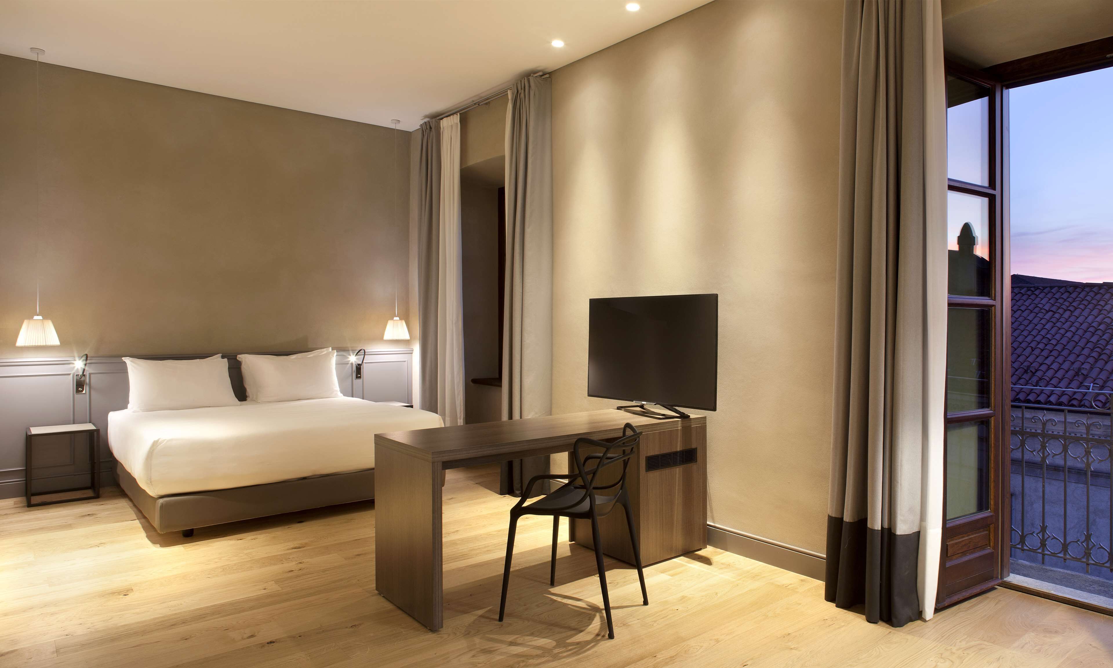 Quarto de hotel com cama, luminárias de cabeceira, escrivaninha com cadeira, televisão de tela plana e uma grande janela com cortinas.