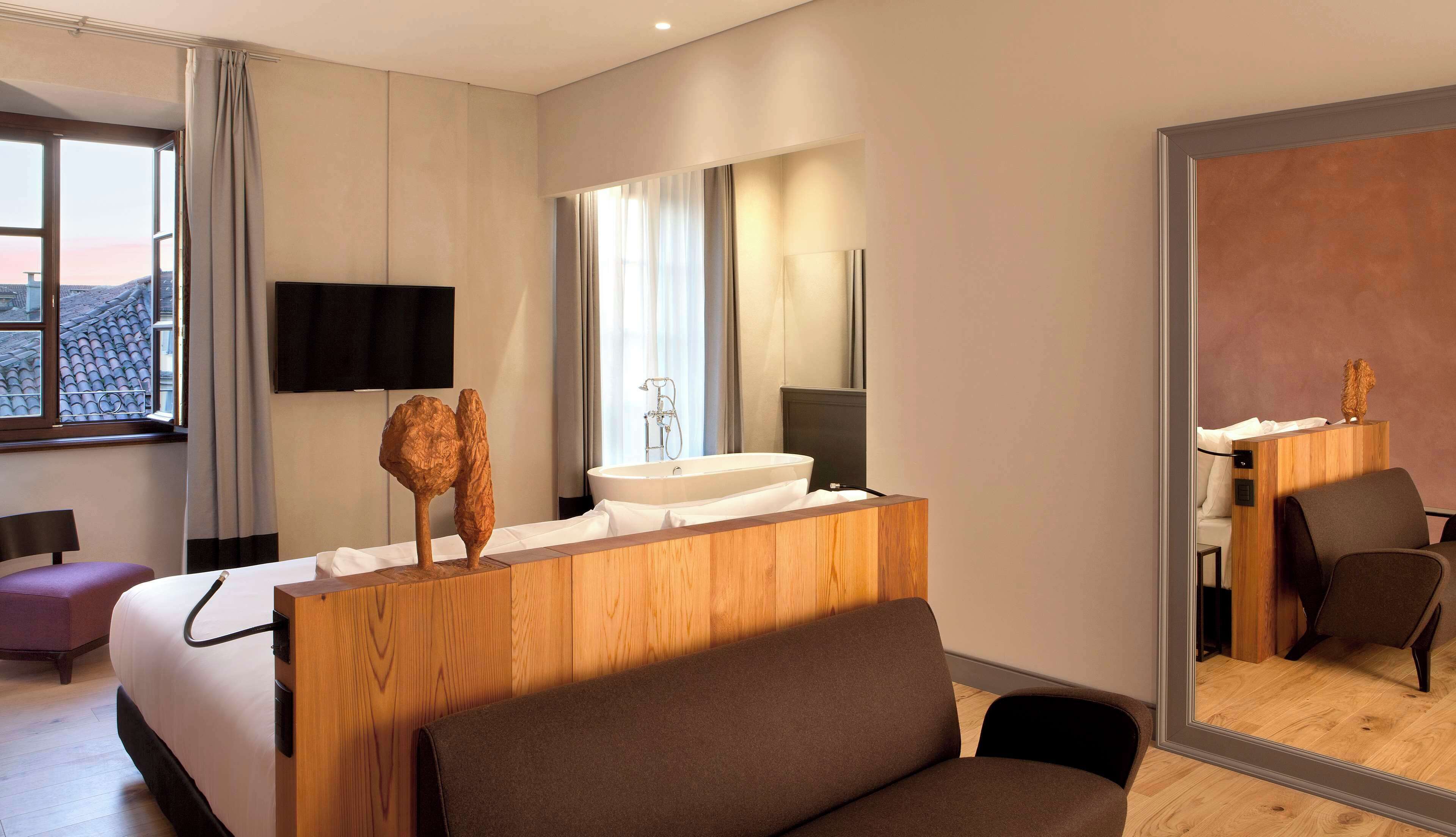 A modern hotel room with a large bed, a wooden vanity with a sink, a flat-screen TV mounted on the wall, and a large mirror reflecting the room.