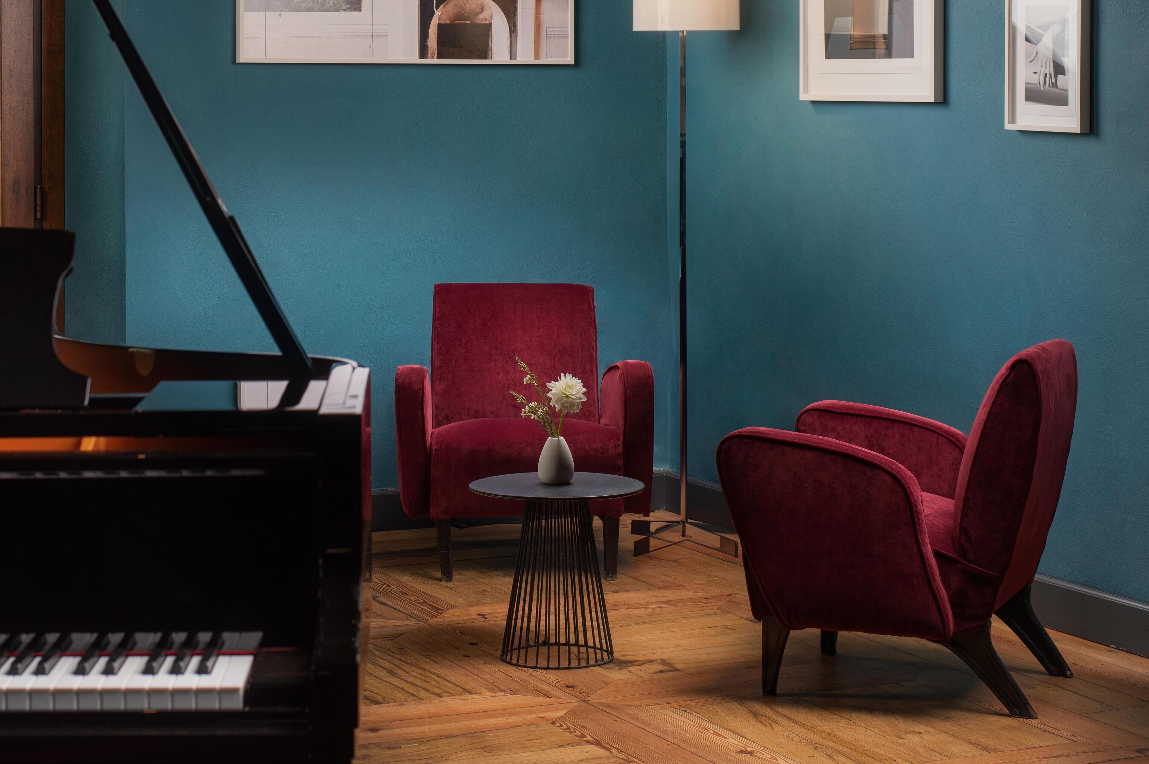 Um quarto com um piano de cauda, duas poltronas de veludo vermelho, uma mesa redonda pequena com um vaso de flores e quadros nas paredes verde-água.