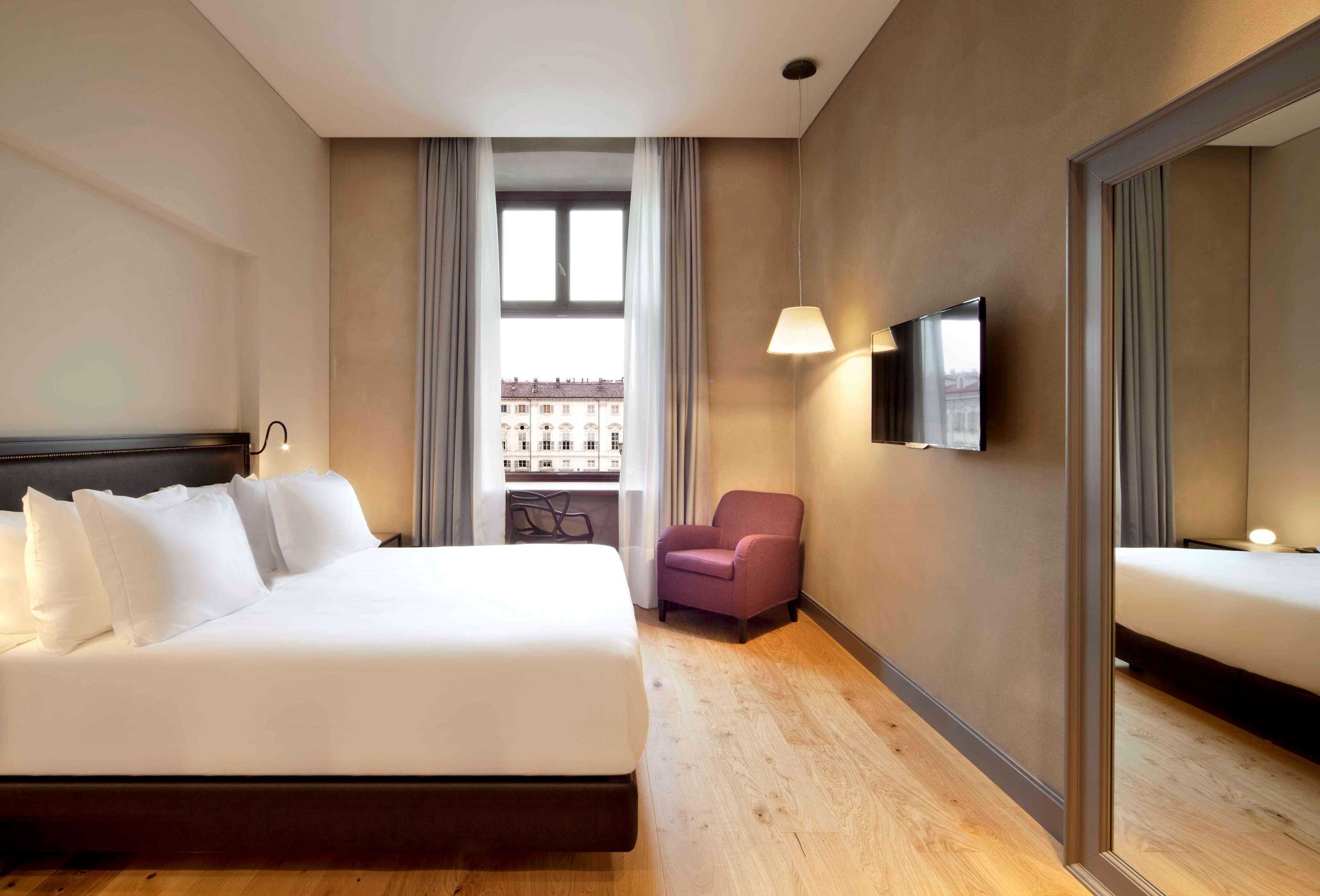 Quarto de hotel com uma cama grande, uma cadeira, uma TV e uma janela com vista para prédios.