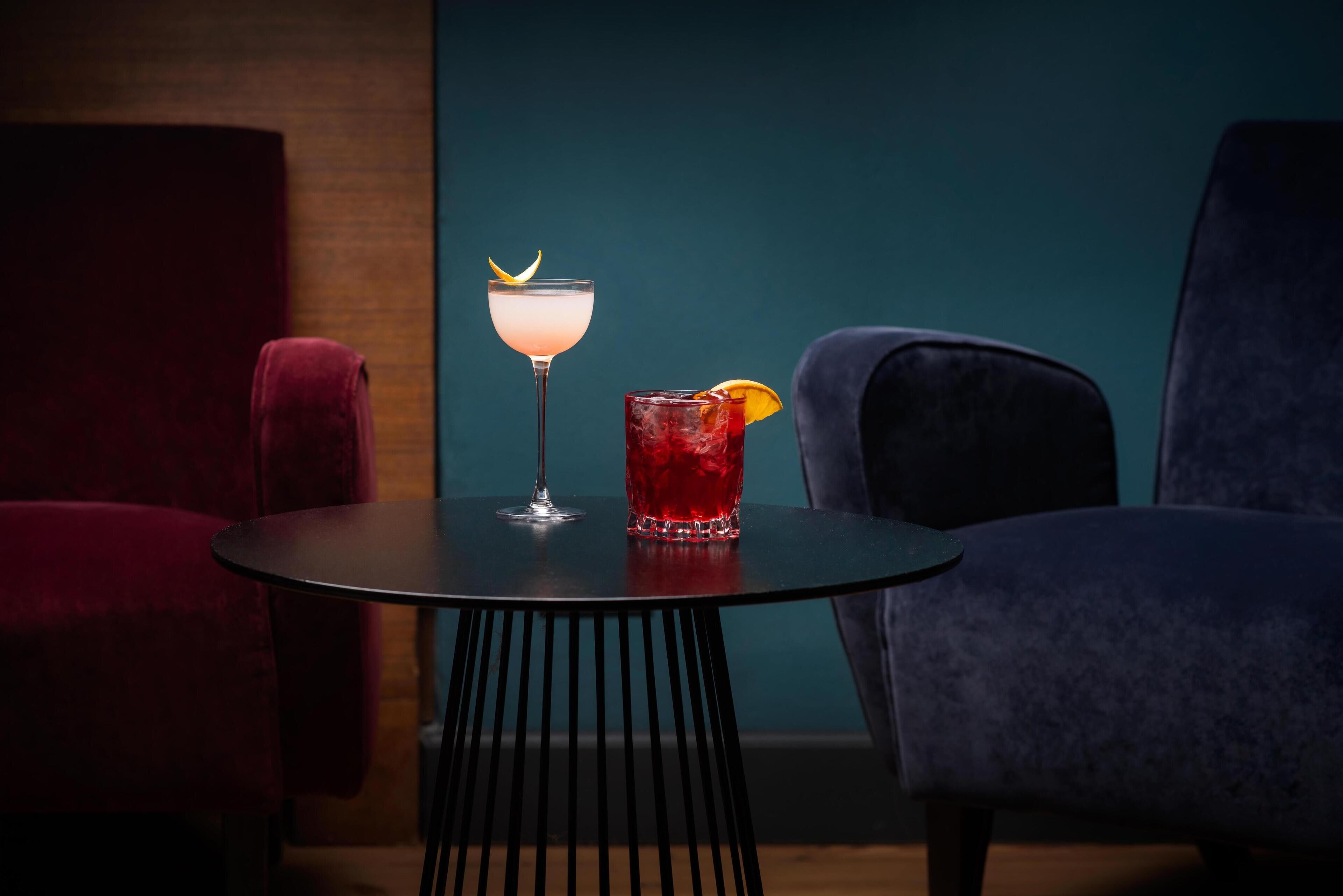 A table with two drinks, a red cocktail and a pale drink with a lemon twist, in a room with dark red and blue chairs.