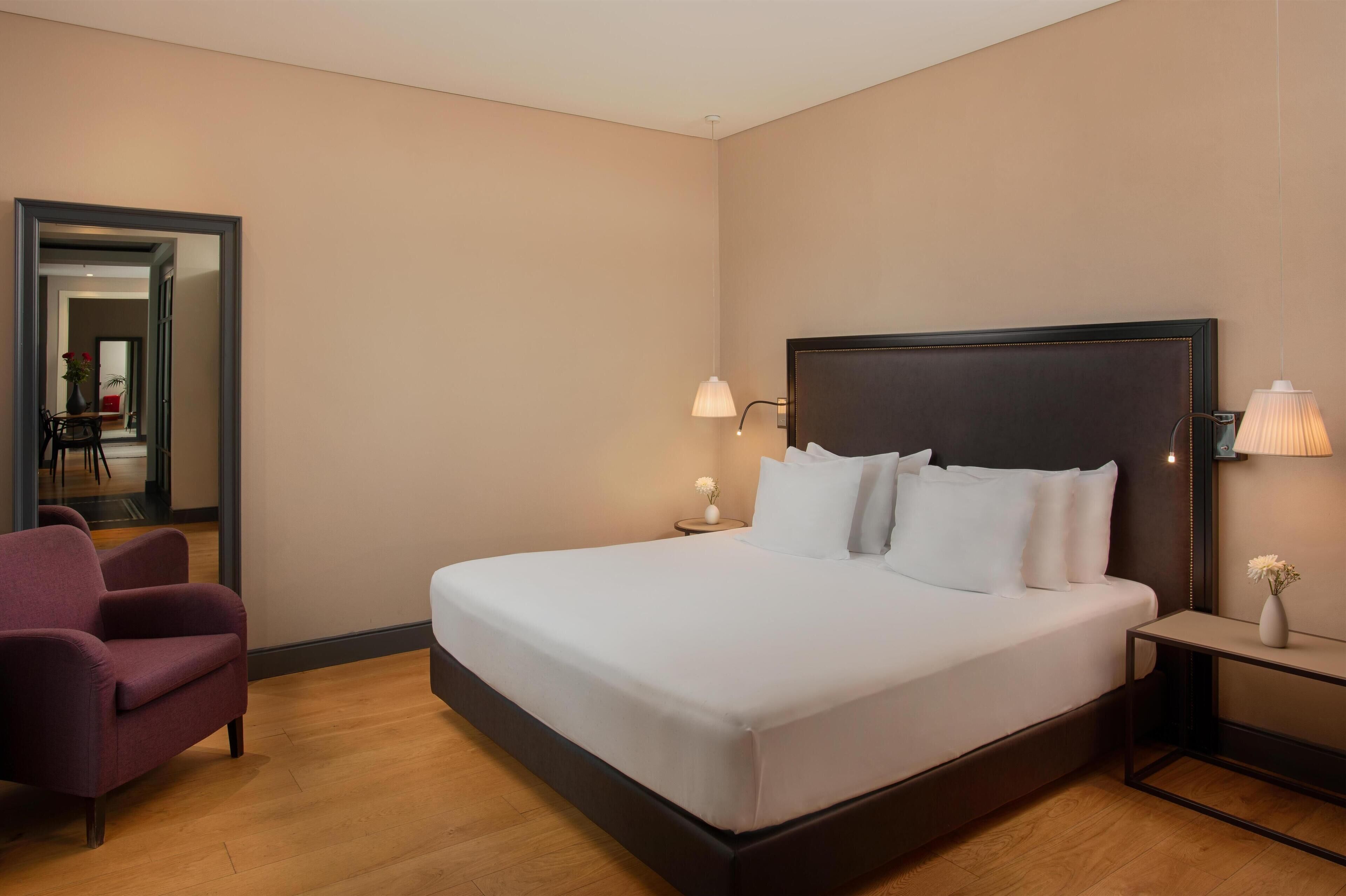 Une chambre d’hôtel bien rangée, avec un grand lit, une chaise, une table de chevet et un miroir.