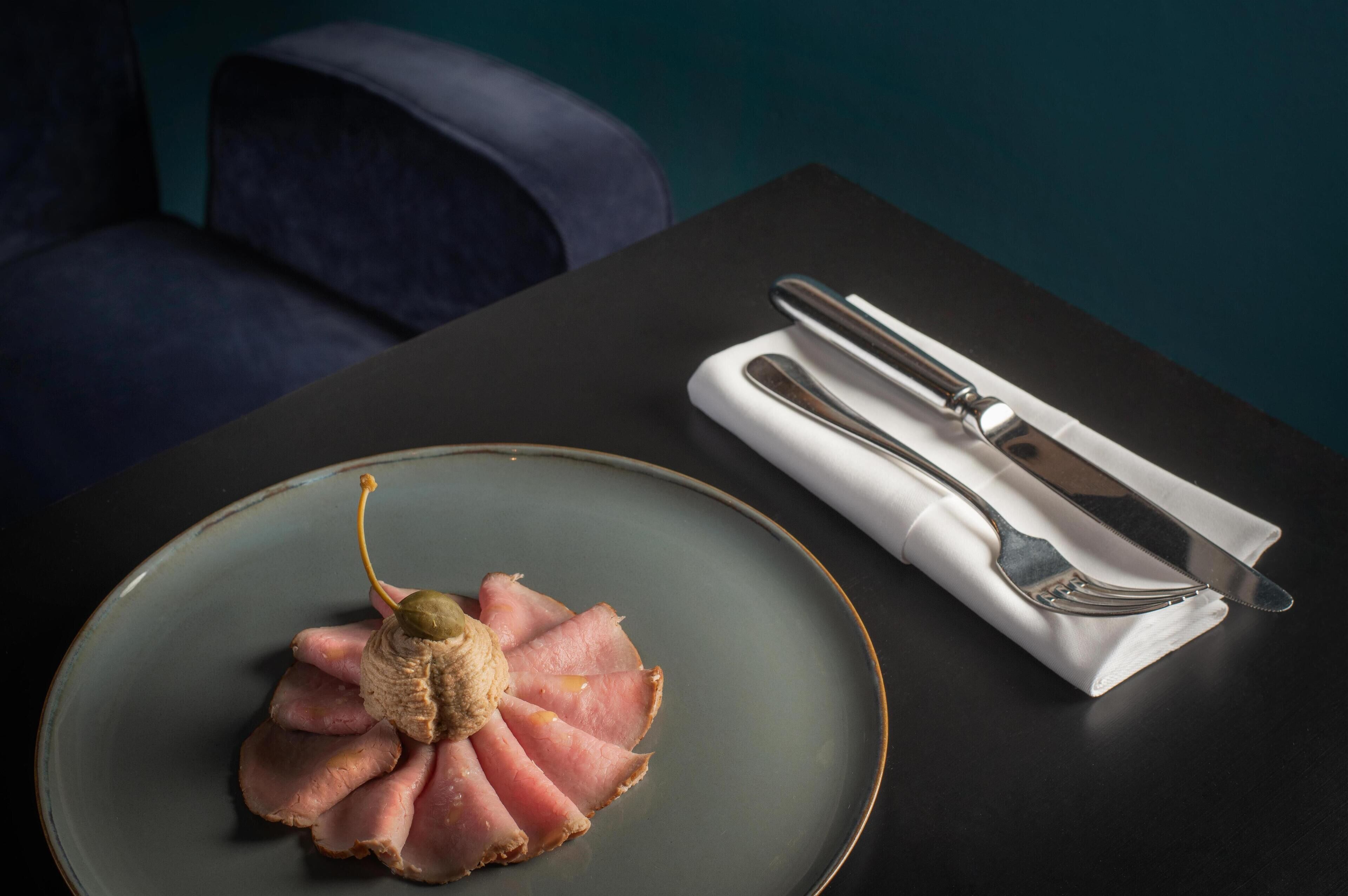 Une assiette de tranches de jambon, agrémentée d’un champignon décoratif, est posée sur une assiette sombre. Une serviette pliée et des couverts complètent la présentation sur la table.