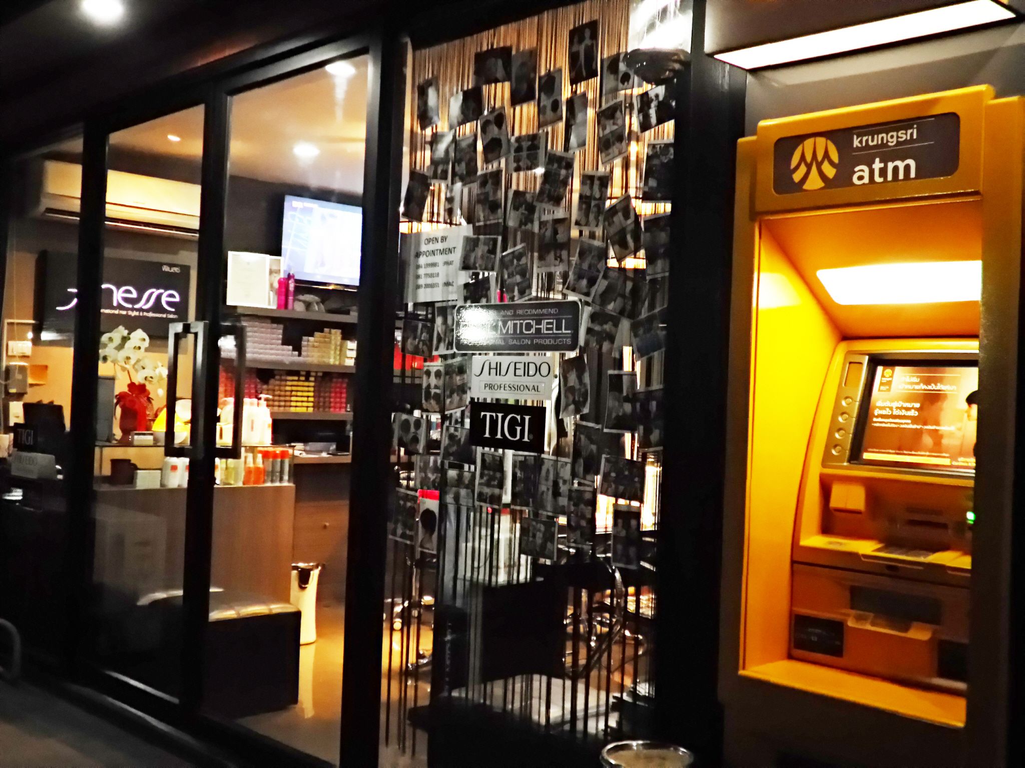 Un distributeur automatique de billets arborant le logo de Krungsri, un salon équipé d’un miroir et proposant divers produits, et un panneau indiquant que le salon est ouvert sur rendez-vous.