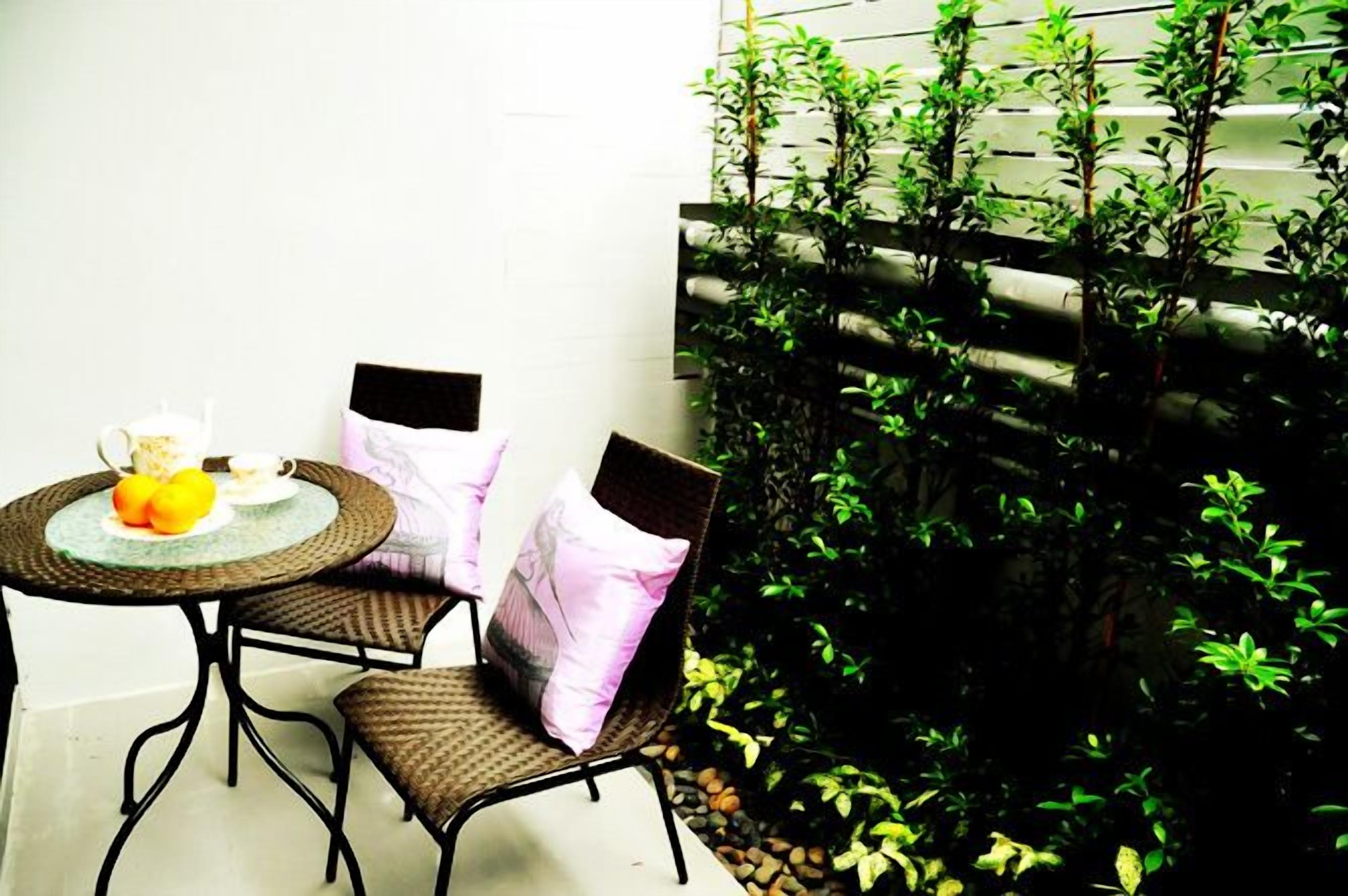 Uma pequena área externa com espaço para sentar, uma mesa redonda e duas cadeiras, parede branca e um painel de madeira coberto por folhagem verde.