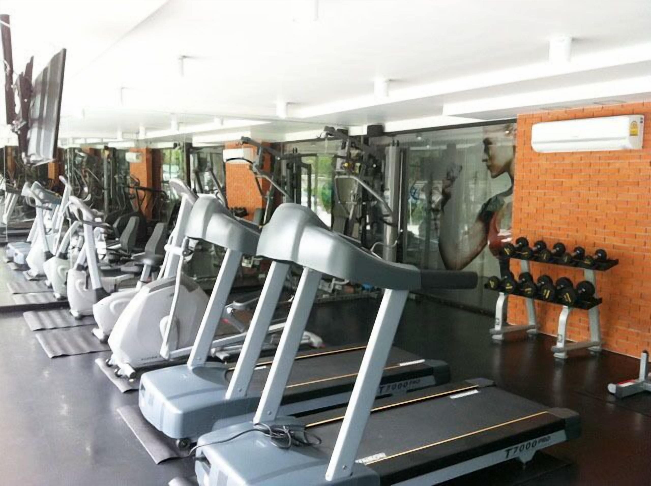 Une rangée de tapis de course dans une salle de sport, avec un mur en briques et une grande fenêtre.