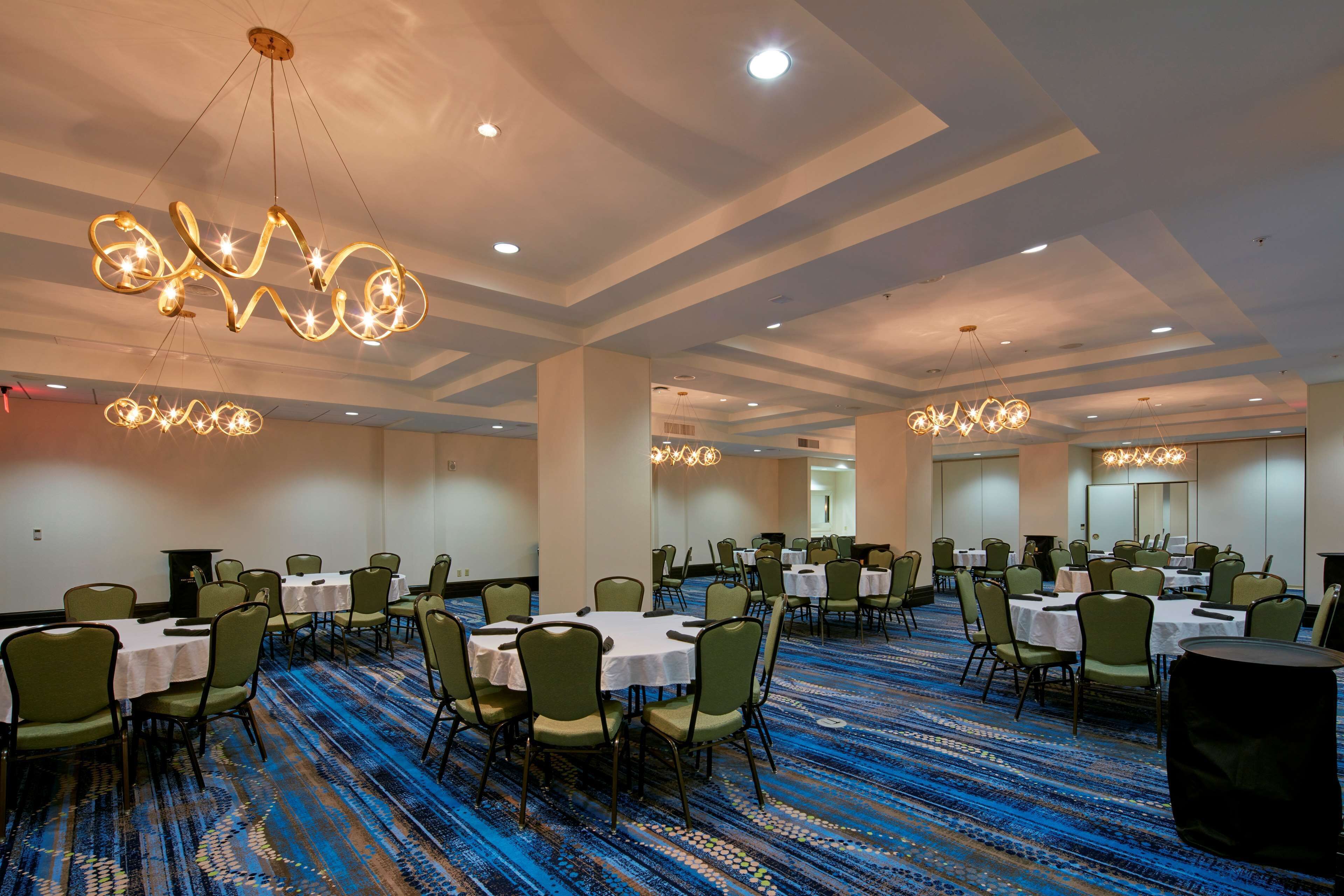 A banquet hall with round tables and green chairs, arranged in rows, with overhead lighting fixtures.