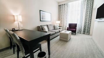 A hotel room with a dining table, a sofa, a chair, a small ottoman, and a TV mounted on the wall.