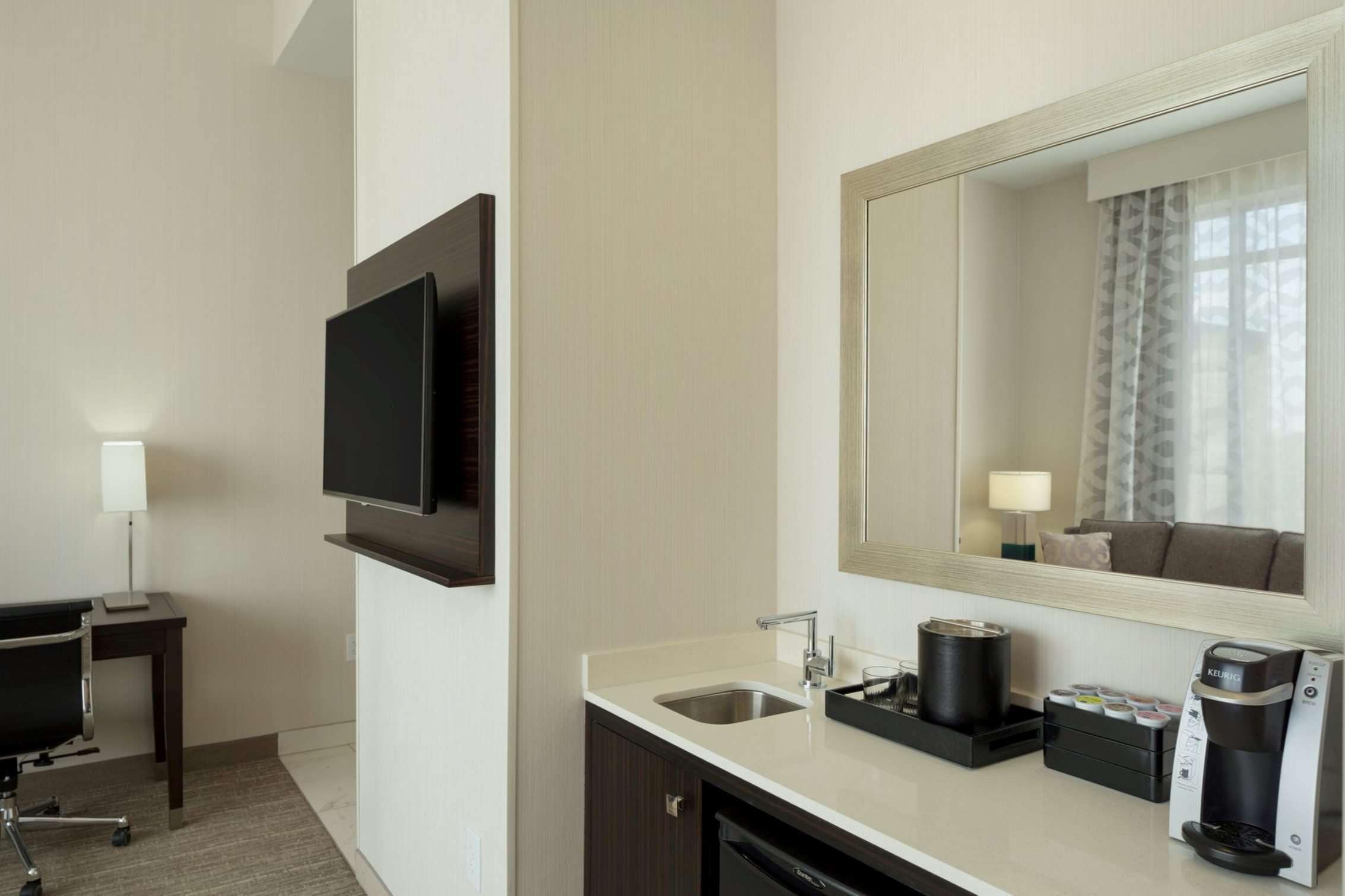 A hotel room with a flat-screen TV mounted on the wall, a sink with a coffee maker, and a mirror above the sink.