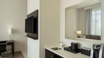 Habitación de hotel con televisor de pantalla plana en la pared, lavamanos con cafetera y espejo sobre el lavamanos.