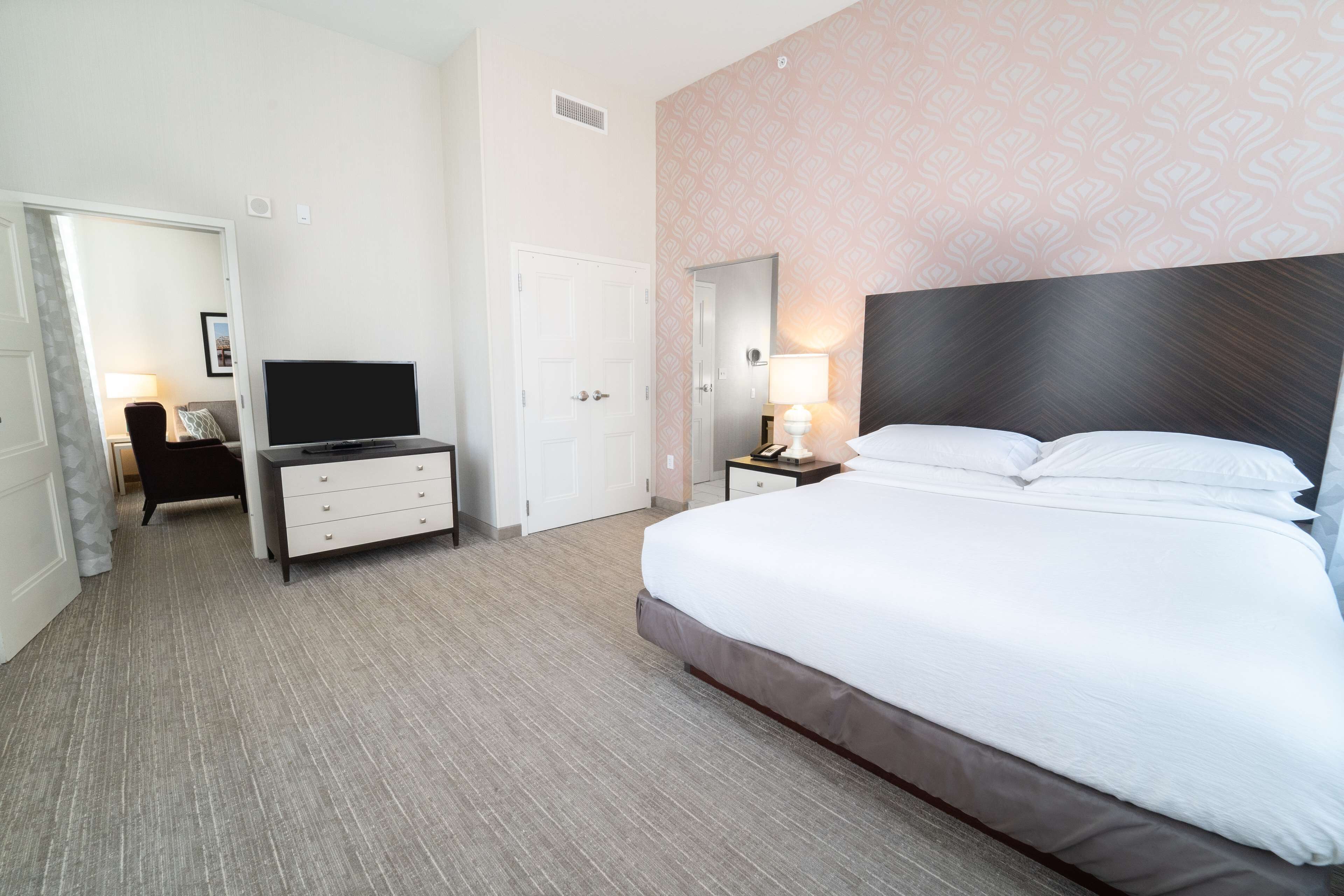Une chambre d’hôtel avec un grand lit, une télévision, un bureau avec des tiroirs, une lampe et une porte donnant sur une autre pièce.