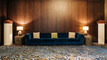 Un sofá azul oscuro con dos almohadas y un cojín decorativo, flanqueado por dos mesitas de noche con lámparas, sobre una alfombra estampada y frente a una pared de madera.