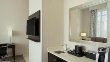 Habitación de hotel con televisor de pantalla plana montado en la pared, un lavamanos con cafetera y un espejo que refleja la habitación.
