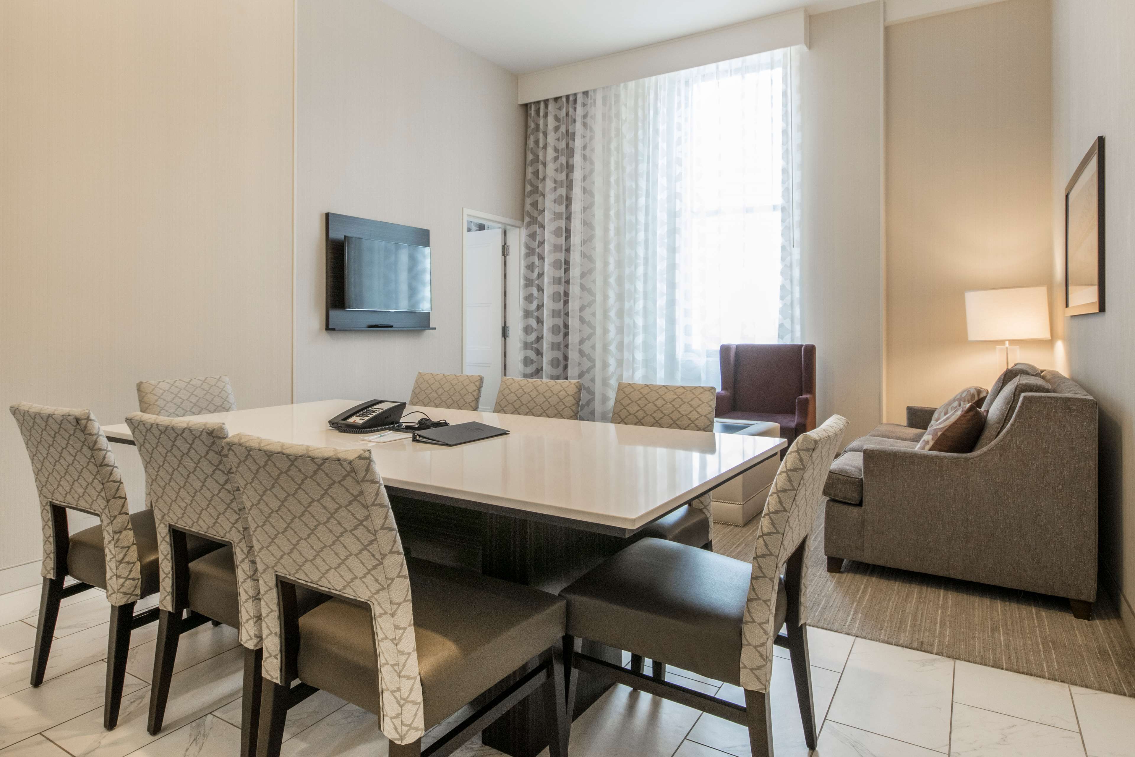 Una habitación de hotel moderna con una mesa de comedor, sillas, un sofá, un televisor y una ventana con cortinas.
