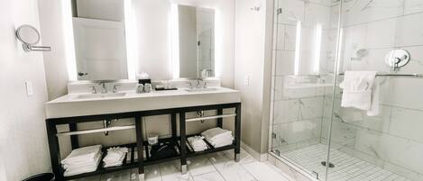 A modern bathroom with a double sink vanity, a glass shower enclosure, and a black trash bin.