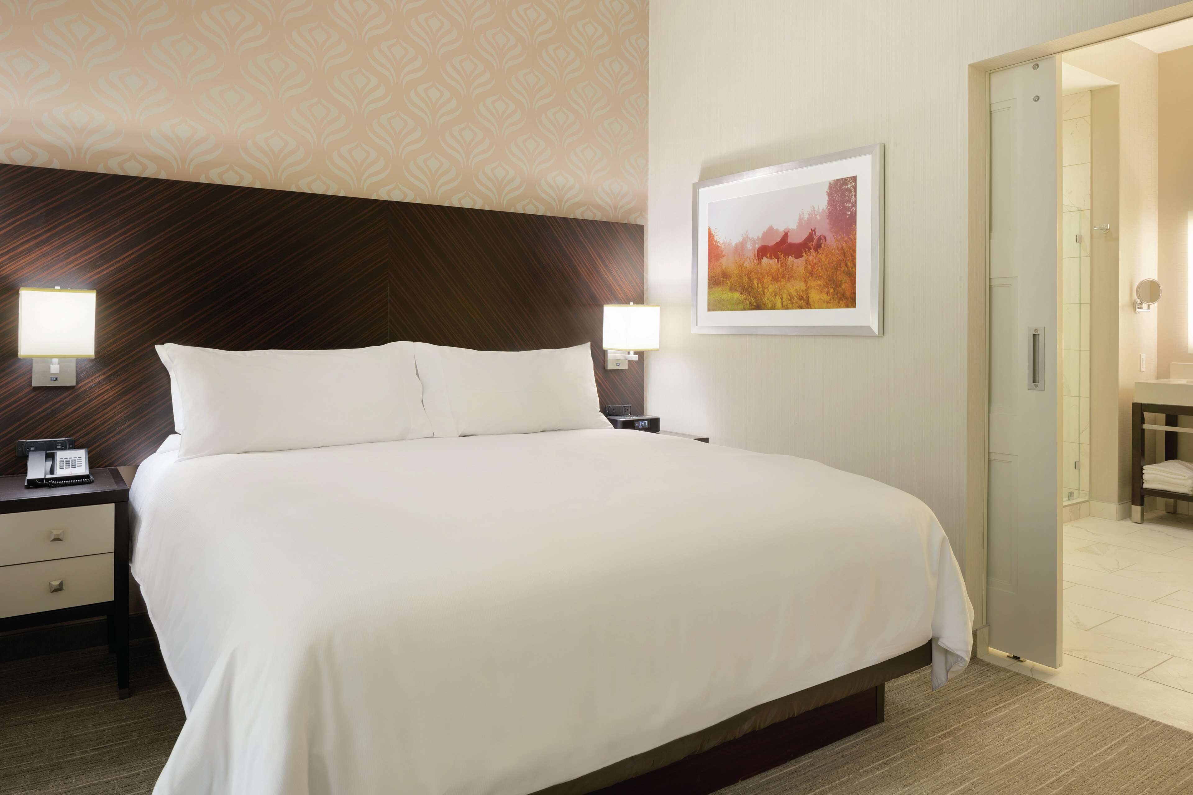 A hotel room with a large bed, bedside lamps, a nightstand, a framed picture on the wall, and a bathroom visible through an open door.
