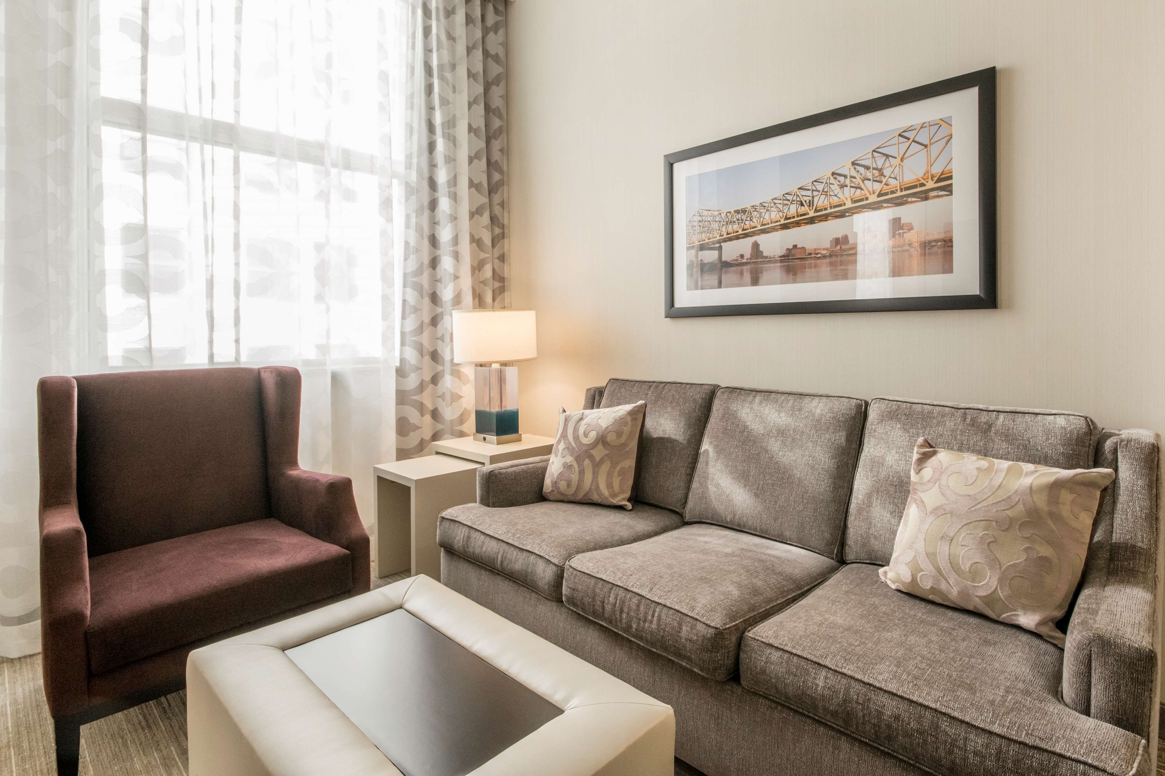 A living room with a brown armchair, a grey sofa, and a white coffee table. There is a lamp on a side table and a framed picture on the wall.