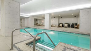 An indoor swimming pool with clear water, surrounded by white marble walls and columns, and furnished with lounge chairs and tables.