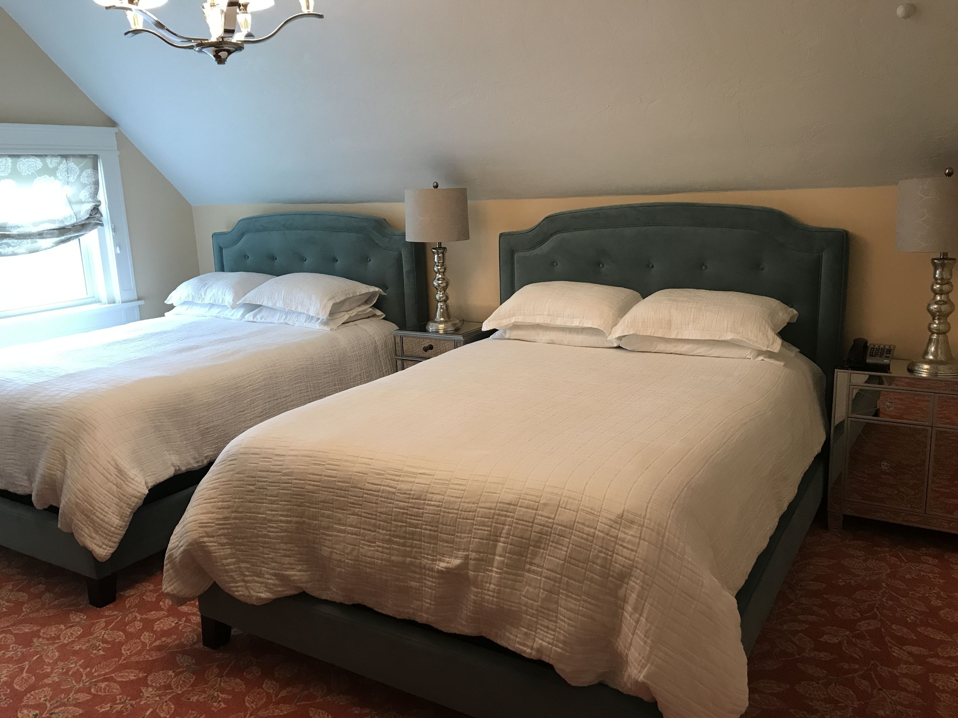 A hotel room with two beds, each with a headboard, bedside tables with lamps, and a chandelier.