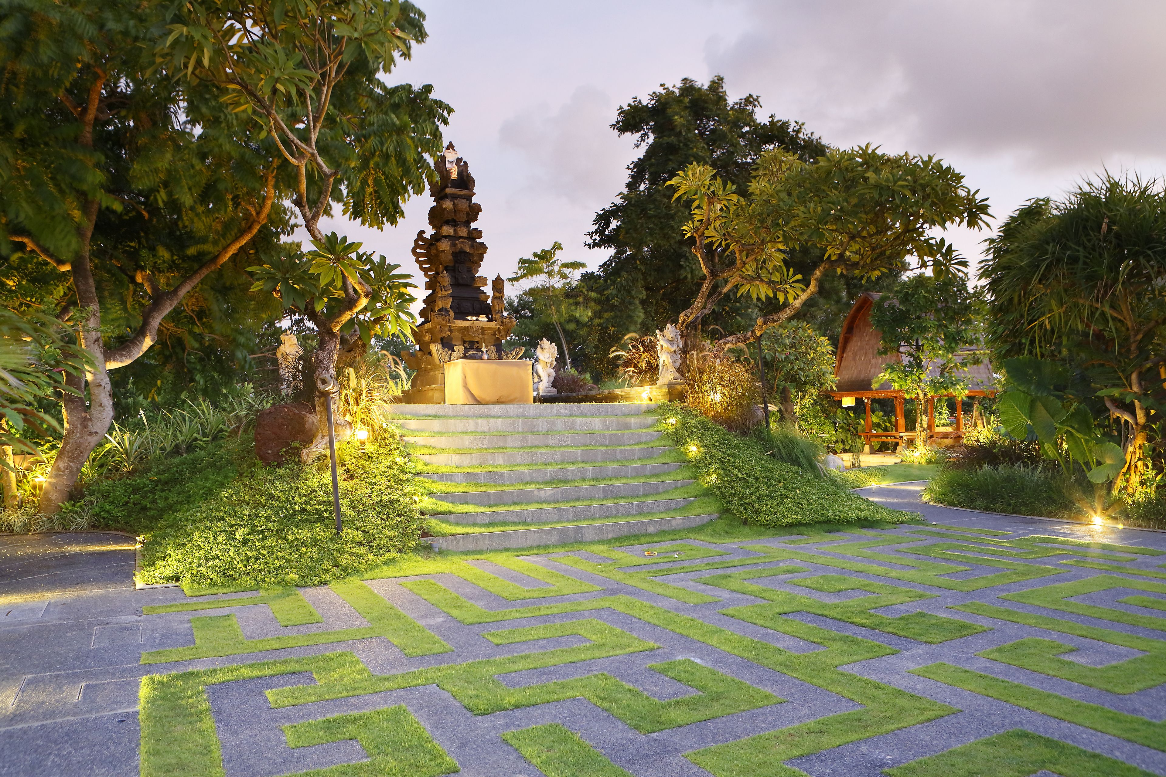 A garden with a stone staircase leading to a pagoda-like structure, surrounded by lush greenery and a patterned stone pathway.