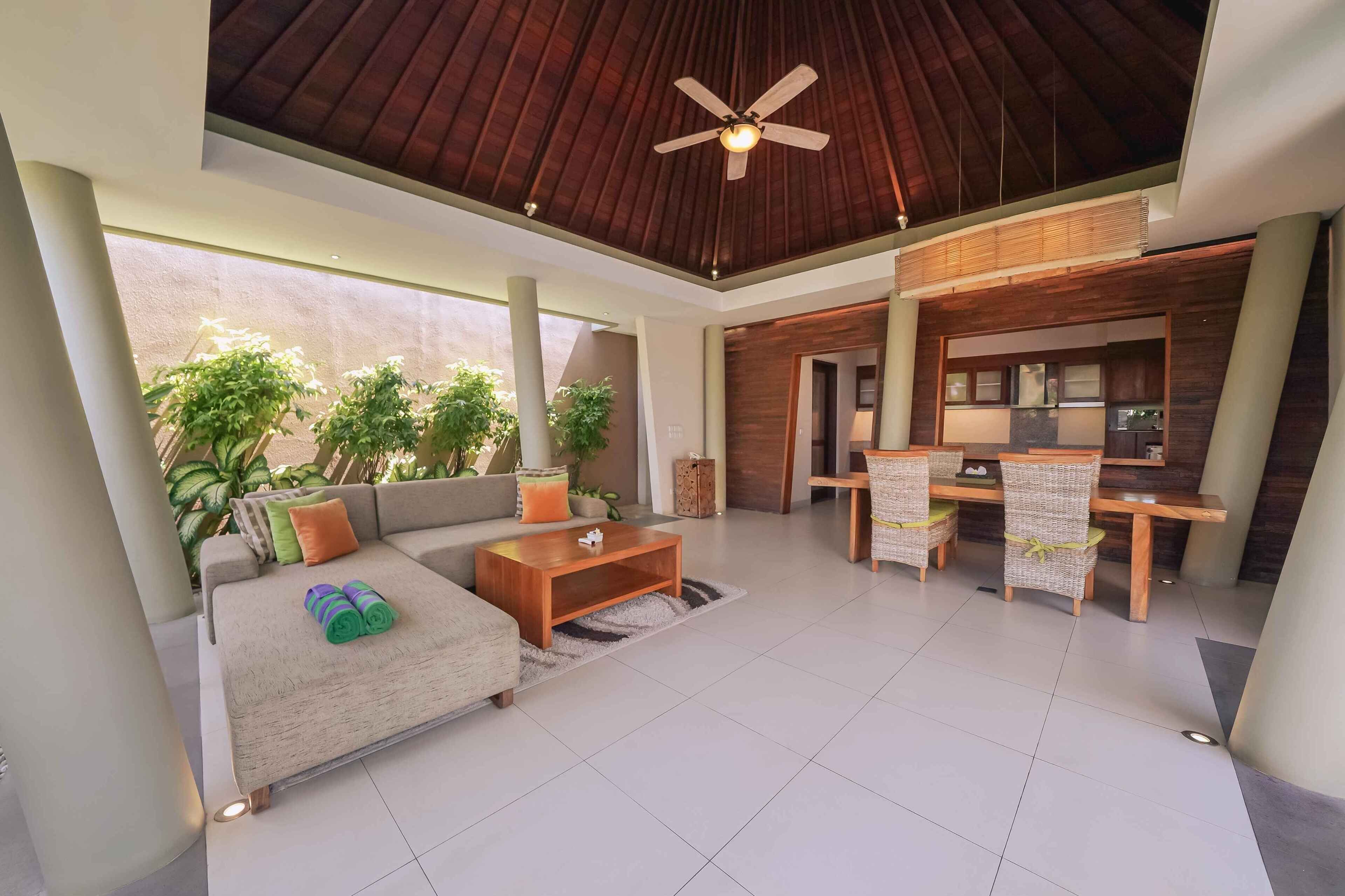 A modern living area with a sofa, coffee table, and dining set. There is a ceiling fan, a view of the outdoors, and a kitchen area visible through an open door.