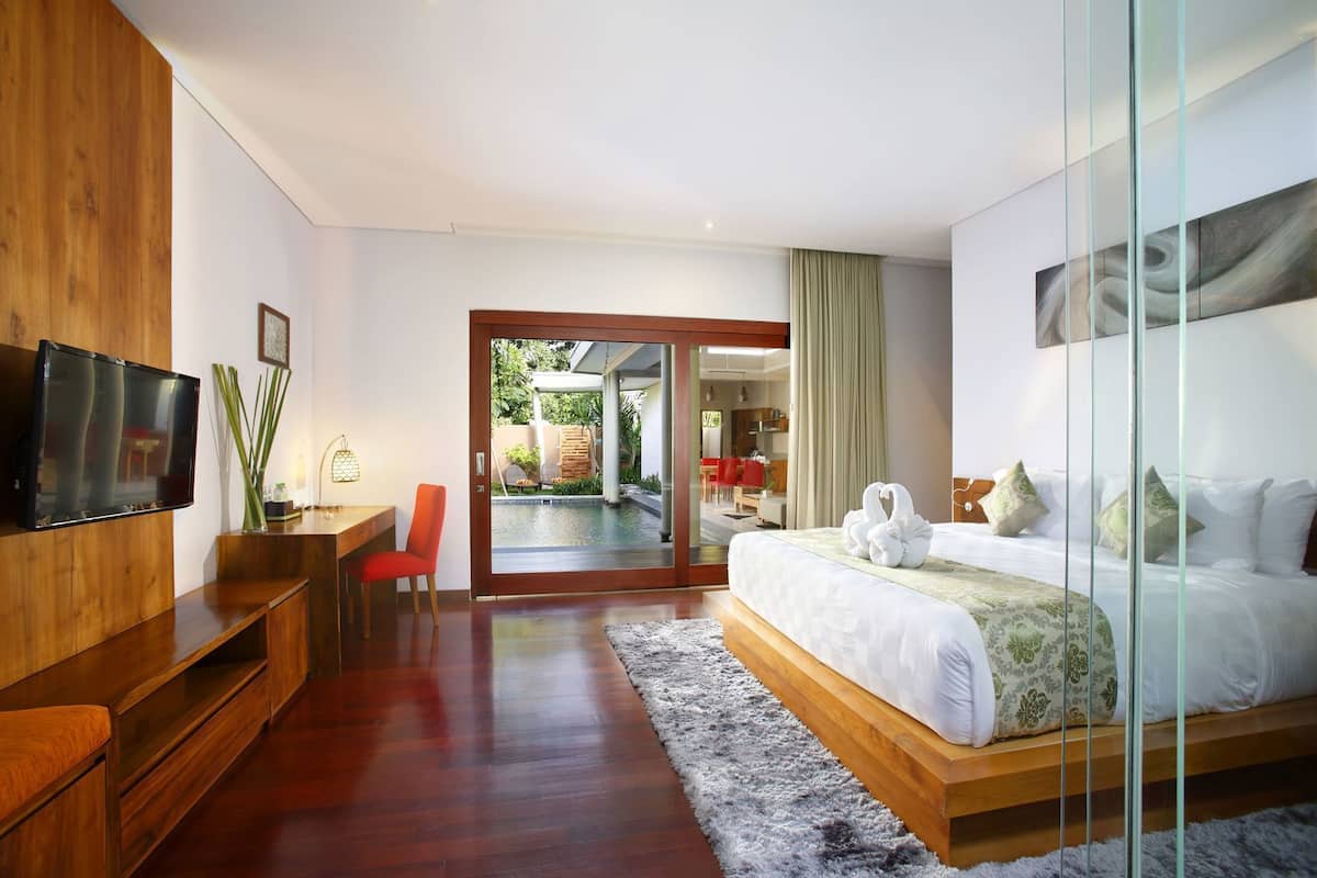 A modern hotel room with a wooden desk, a flat-screen TV, a bed with white bedding, and a glass door leading to a pool area.