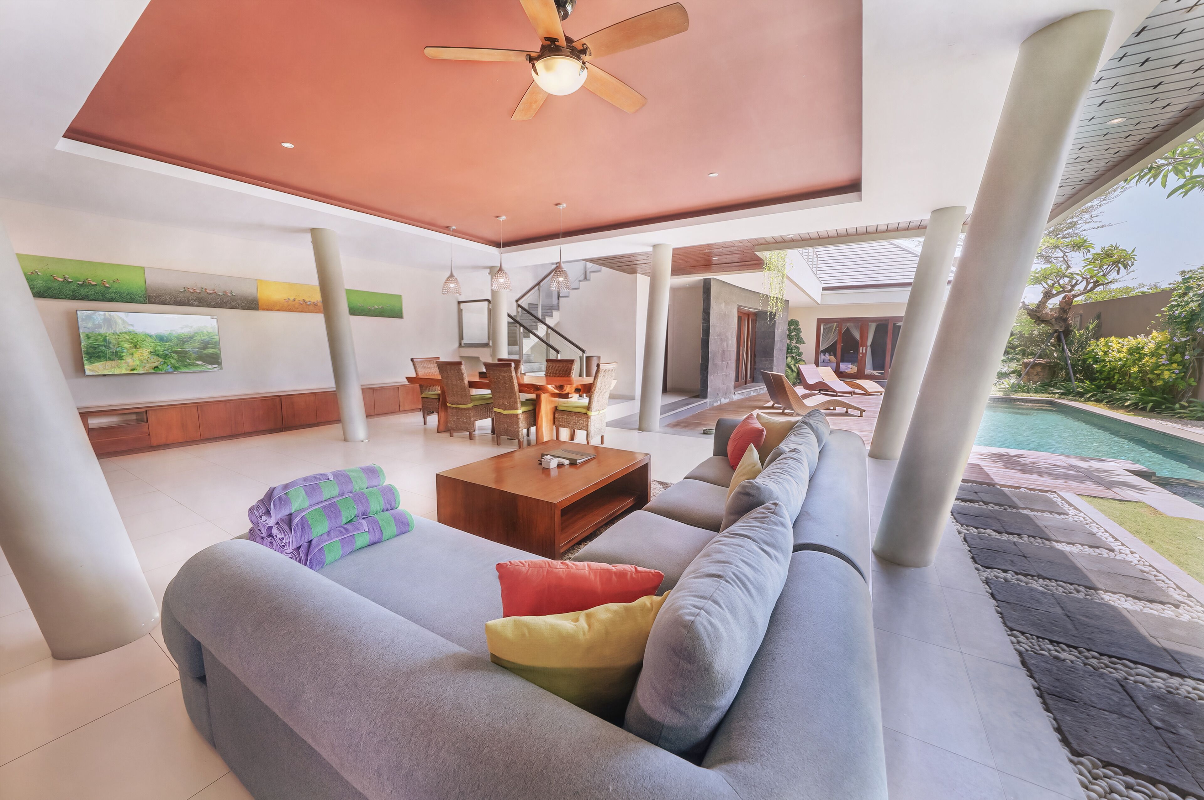 A spacious living area with a sofa, coffee table, and a dining table with chairs. There is a ceiling fan, a TV mounted on the wall, and a staircase leading to another level.
