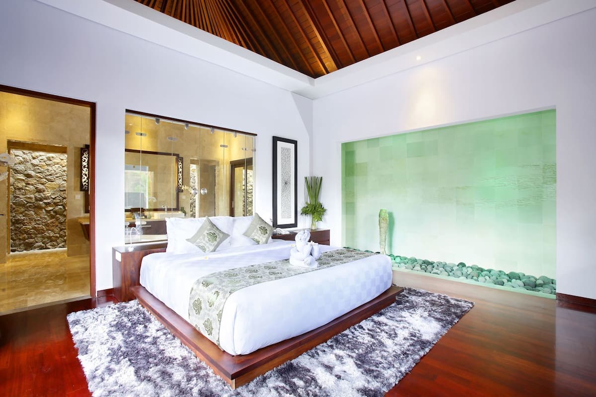 A modern bedroom with a large bed, a stone wall, a green wall with a water feature, and a wooden ceiling.
