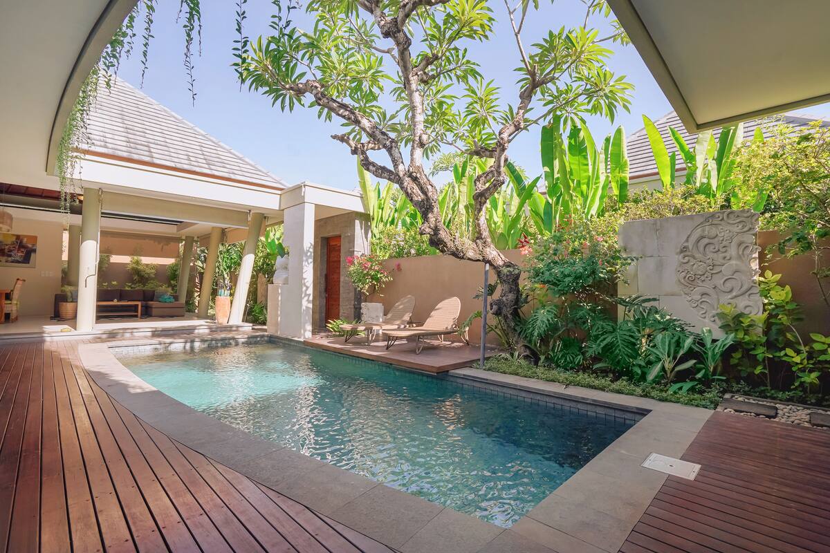 A pool area with wooden decking, lounge chairs, and a covered seating area.