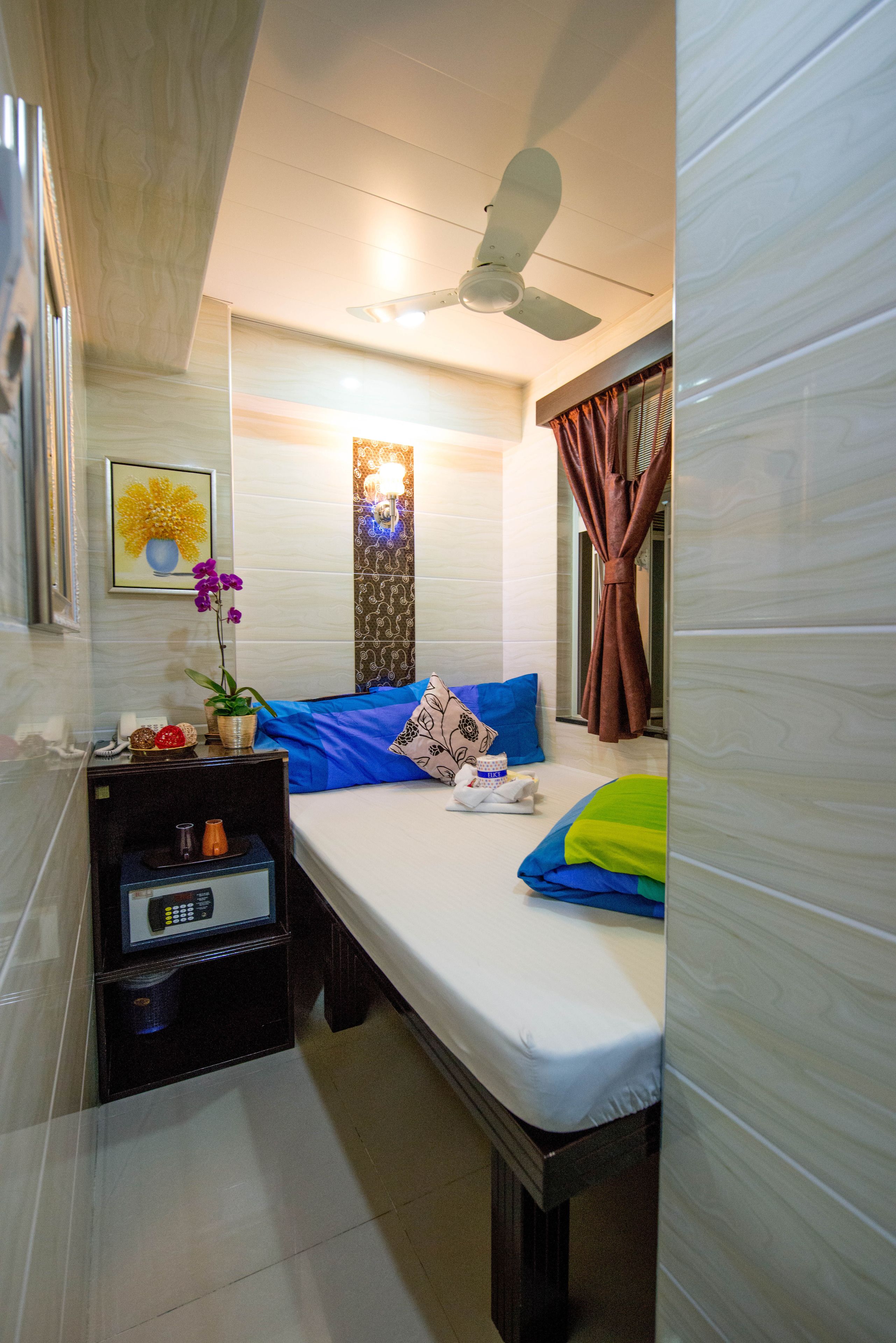 A compact hotel room with a bed, a bedside table with a lamp, a ceiling fan, and a window with curtains.