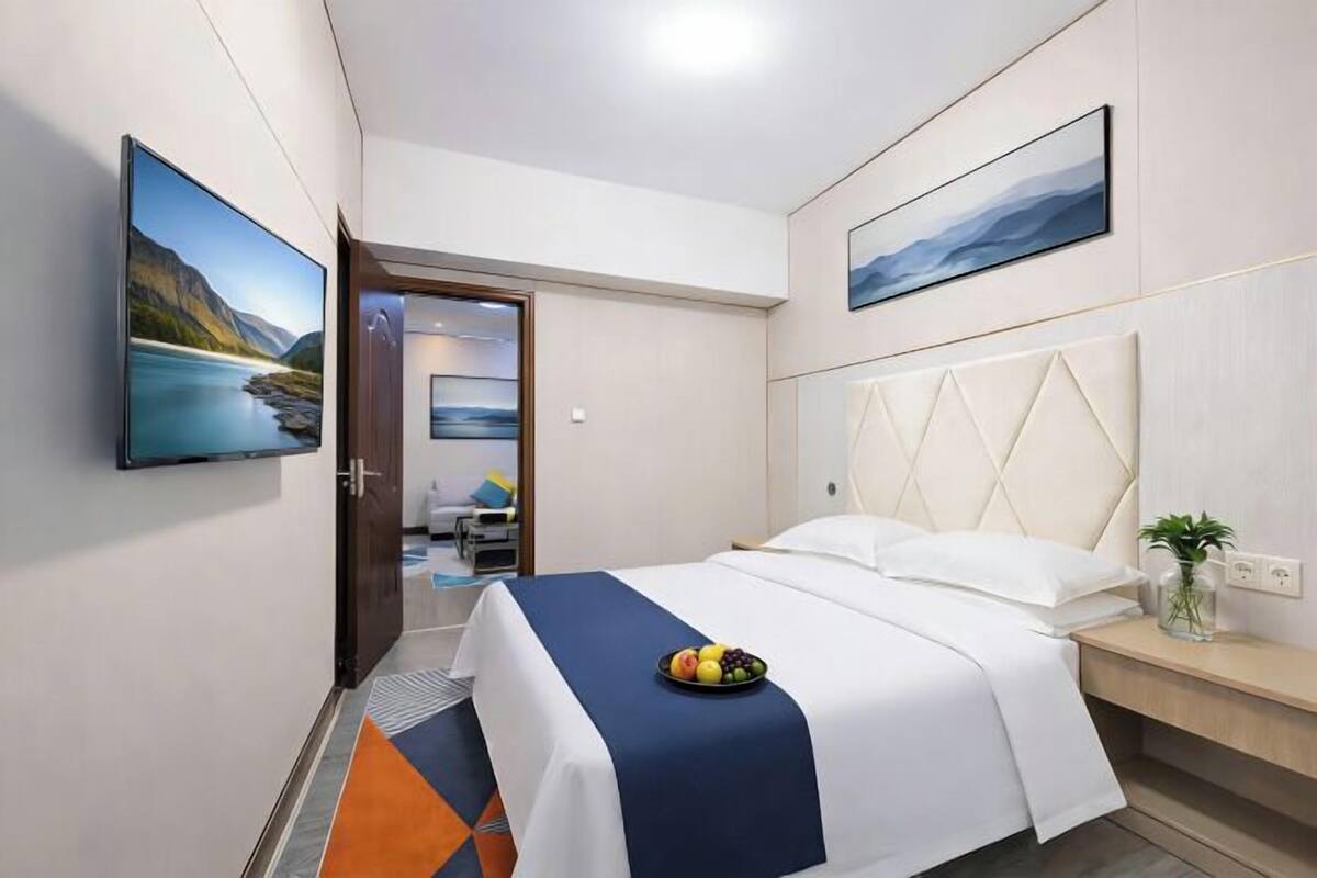 Een hotelkamer met een bed, een nachtkastje met een plant, een schaal fruit en twee schilderijen aan de muur.