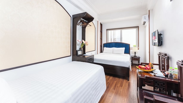 A hotel room with two beds, a desk with a chair, a small table with a basket of fruit, a mirror, a TV, and a window. - Little Hanoi Hotel (Hanoi)