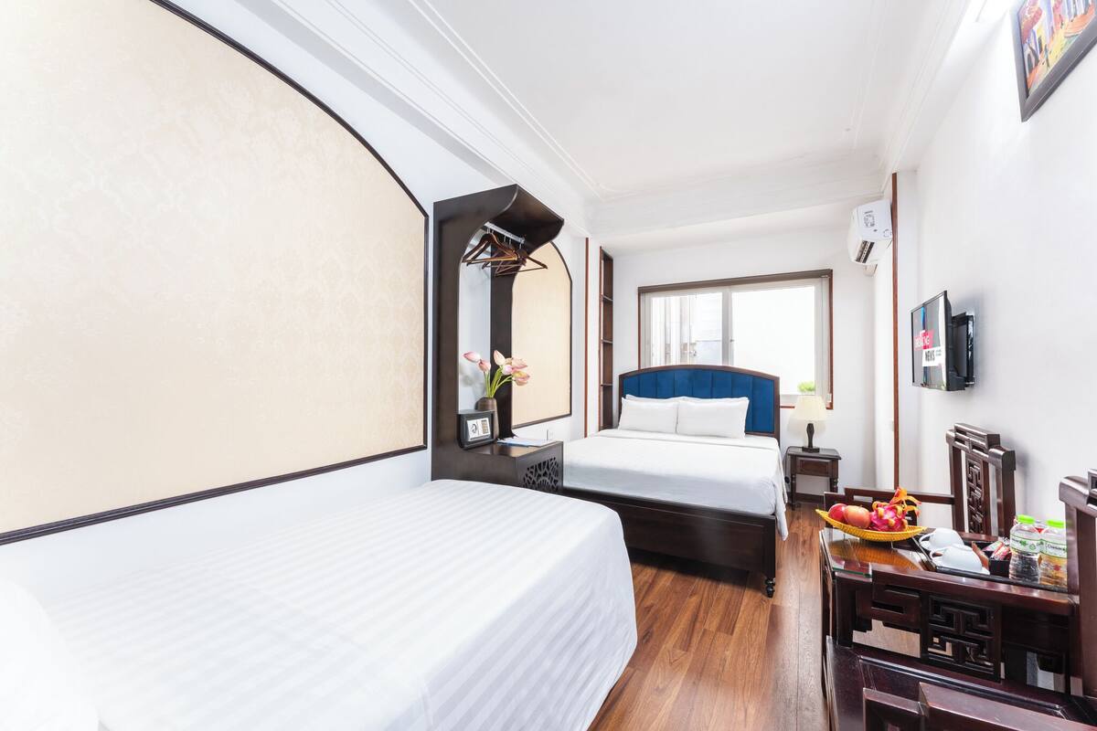 A hotel room with two beds, a desk with a chair, a small table with a basket of fruit, a mirror, a TV, and a window.