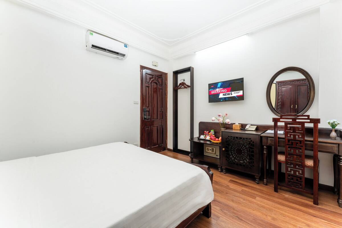 A hotel room with a bed, a desk with a chair, a TV mounted on the wall, and a wooden wardrobe.