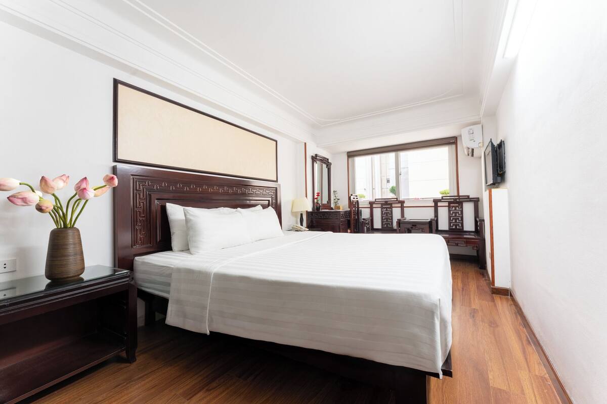 A hotel room with a large bed, a bedside table with a vase of flowers, a wooden headboard, a mirror, and a television on the wall.
