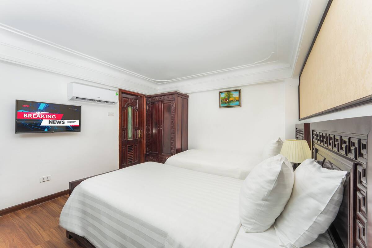 A hotel room with two beds, a TV on the wall, a painting, and a wooden wardrobe.