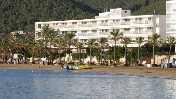 A hotel with multiple floors, a beach in the foreground, and palm trees. - Arenal (Sant Antoni de Portmany)