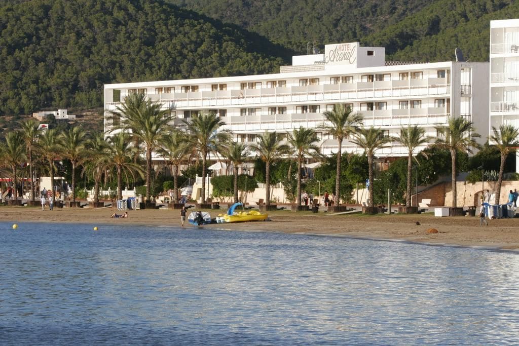 A hotel with multiple floors, a beach in the foreground, and palm trees.