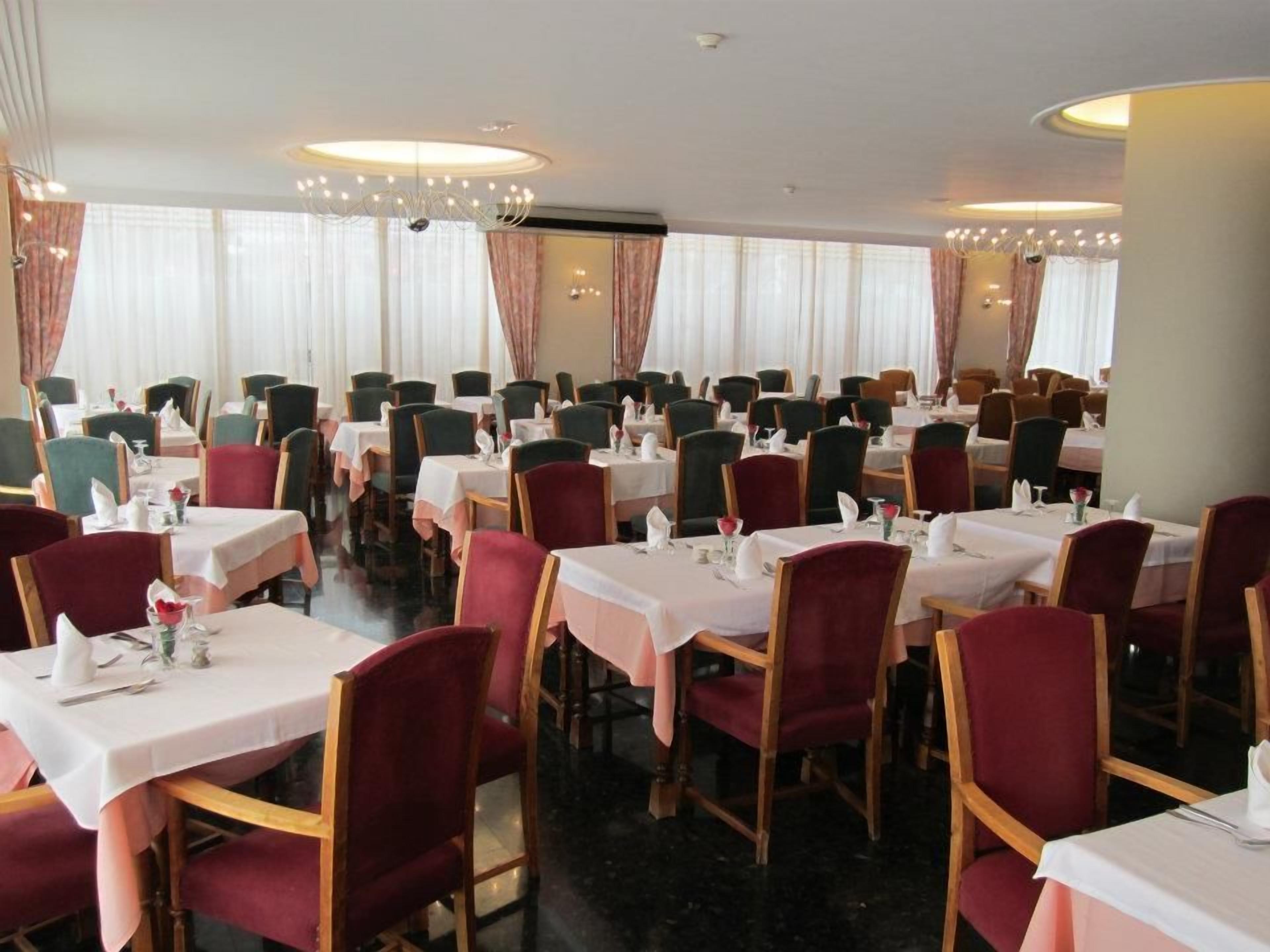 A dining area with multiple tables and chairs, arranged in rows, with a chandelier and curtains visible.