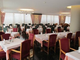 Un comedor con varias mesas y sillas dispuestas en filas, con una araña y cortinas a la vista.