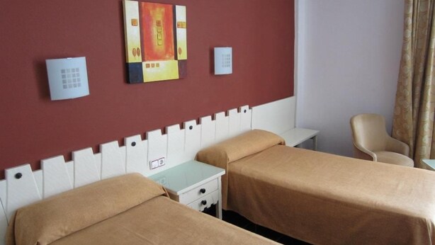 Ein Hotelzimmer mit zwei Betten, einem weißen Kopfteil, einem kleinen Nachttisch, einem Sessel und einem an der Wand befestigten Thermostat.