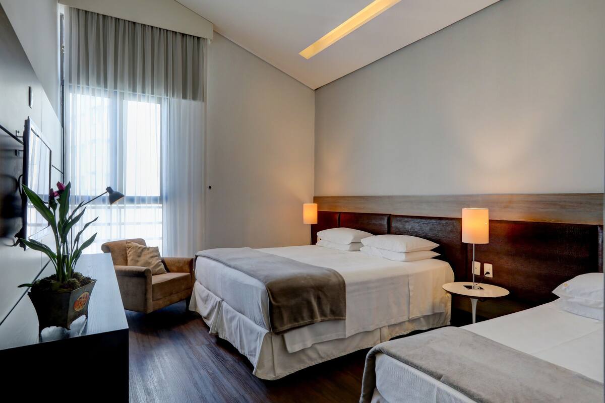 Ein Hotelzimmer mit zwei Betten, einem Schreibtisch, einem Stuhl, einer Topfpflanze und einem fenster mit Vorhängen.
