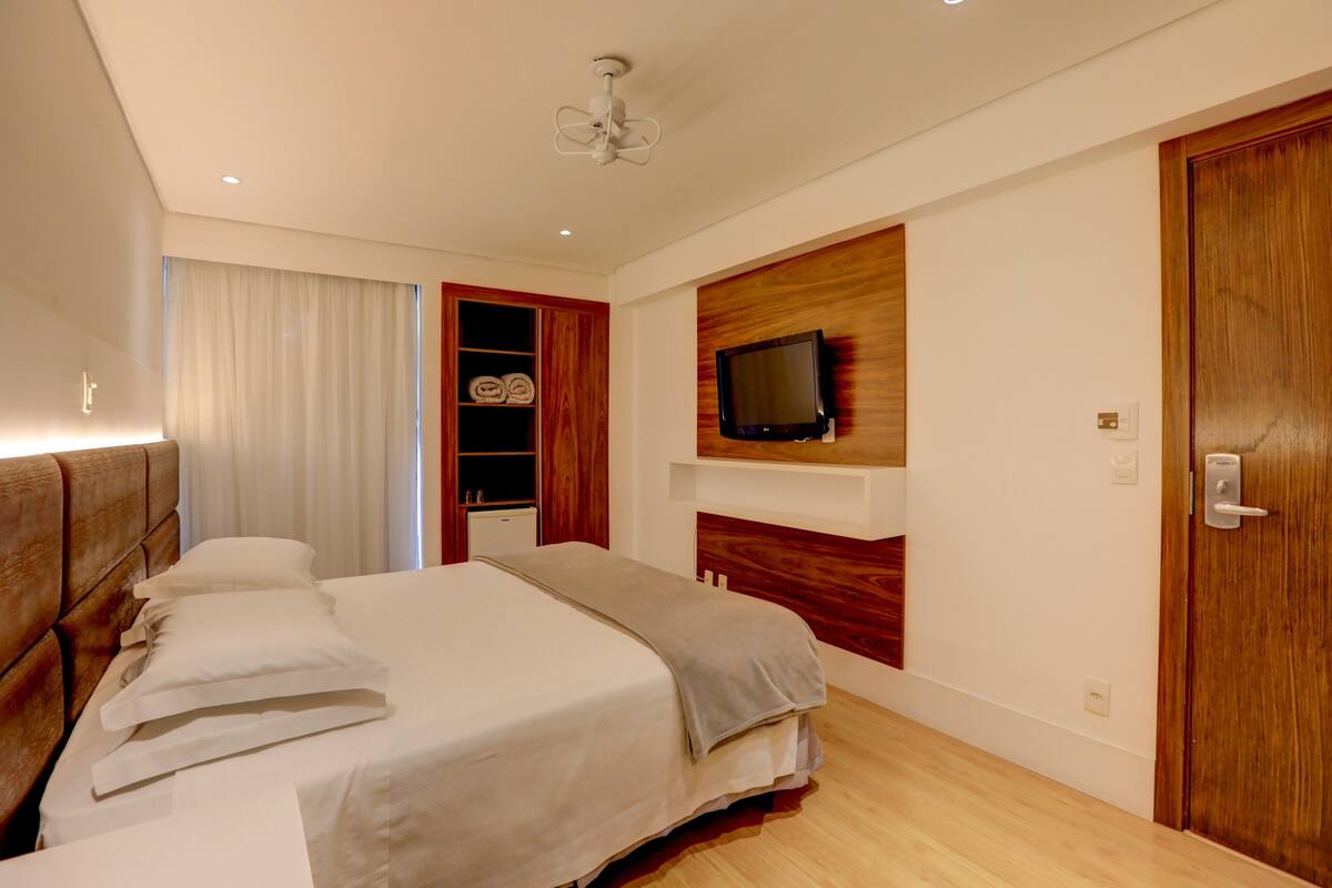 Ein Hotelzimmer mit einem großen Bett, einem an der Wand montierten Fernseher, einem Holzschrank und einer Tür mit Griff.