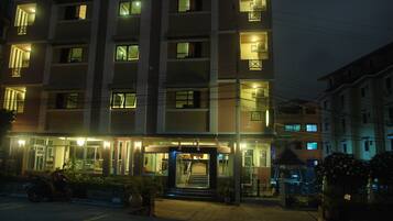 A multi-story hotel with a central entrance, illuminated windows, and a parking area with a motorcycle.