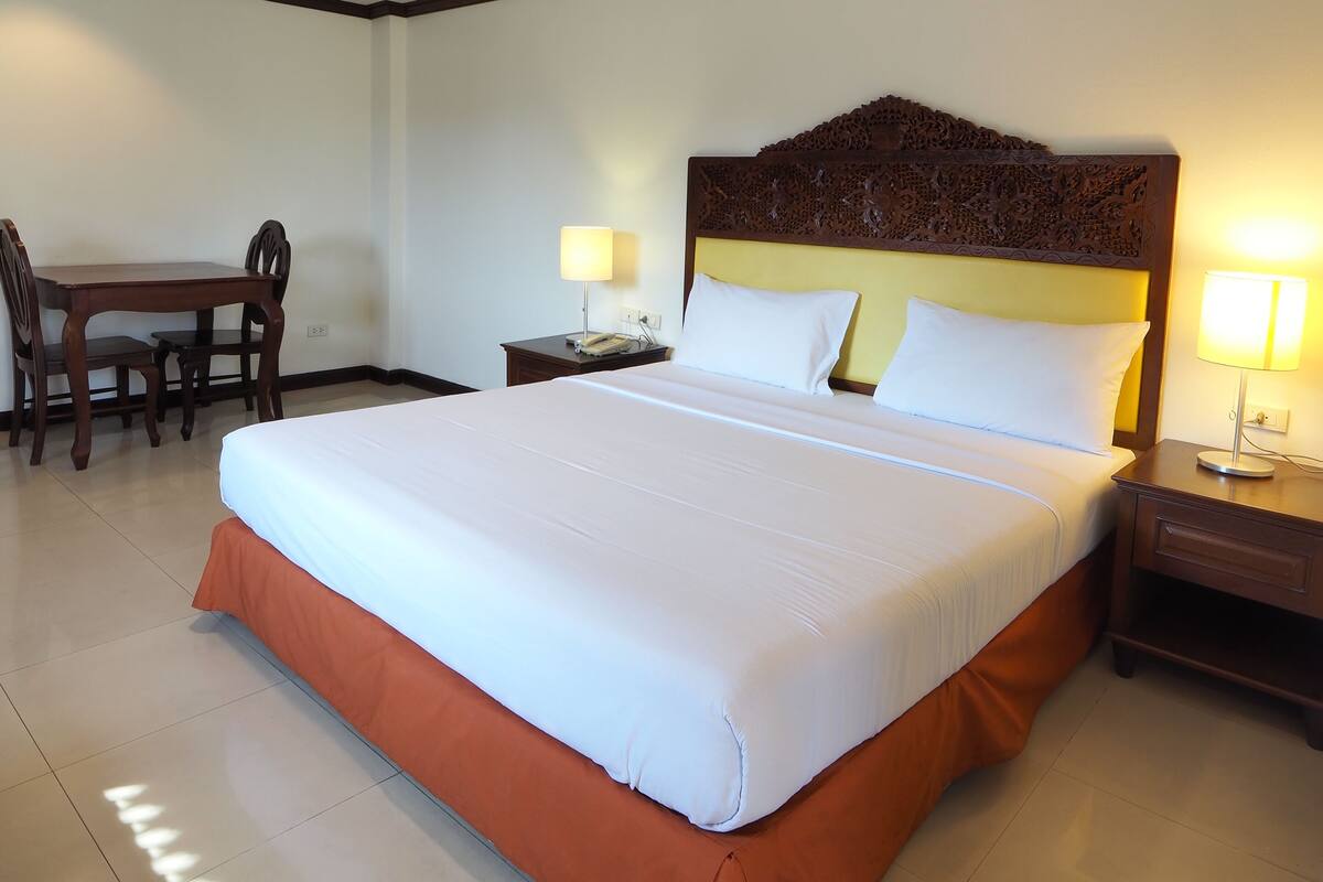 A hotel room with a large bed, a desk with a chair, two bedside lamps, and a wooden headboard.