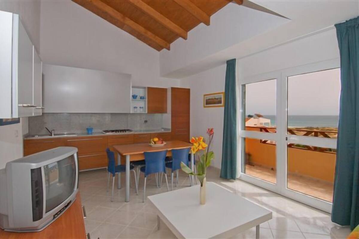A compact kitchen with modern appliances, a dining area with a table and chairs, and a large sliding glass door leading to a balcony with a view of the beach.